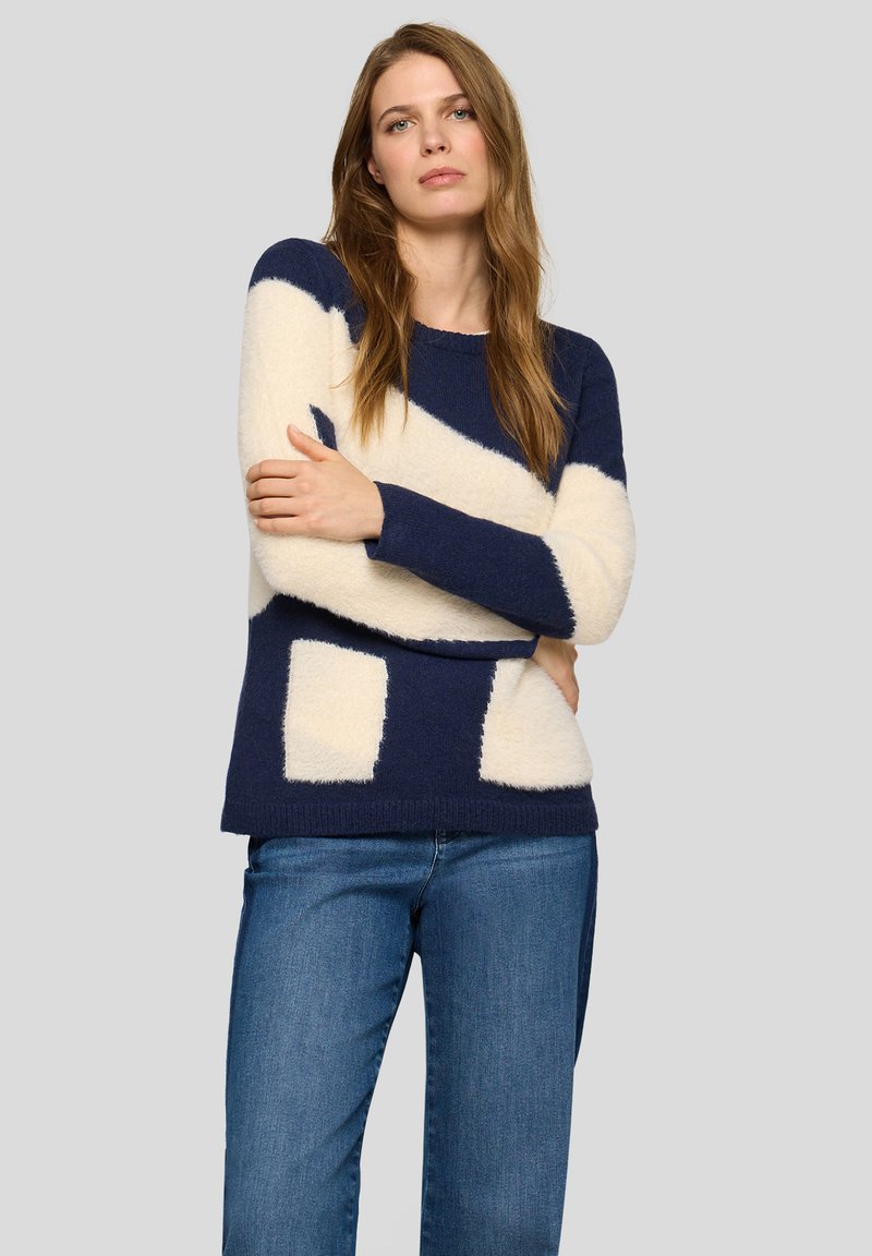 Frau mit langen Haaren, die einen marineblauen und cremefarbigen Pullover im Blockdesign und blaue Jeans trägt, mit verschränkten Armen vor einem grauen Hintergrund steht.