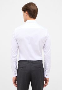 Camicia bianca con maniche lunghe, bottoni ai polsini e una texture liscia, abbinata a pantaloni grigi con due tasche posteriori e bottoni.