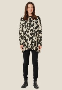Beżowy sweter tunika z dzianiny z czarnym kwiatowym wzorem, długie rękawy, boczne rozcięcia, zestawiony z czarnymi jeansami slim fit i czarnymi botkami za kostkę.