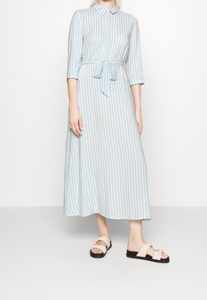 Vrouw draagt een lichtblauwe en witte midi-jurk met verticale strepen, driekwartmouwen en beige slip-on sandalen met zwarte zolen.