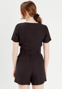 Combinaison noire à manches courtes, avec une taille ajustée et une ceinture, présentant une texture lisse et des poches arrière. Design simple et minimal.