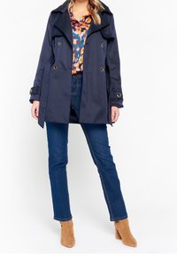 Marinblå trenchcoat med dubbelknäppt design, guldknappar och bred krage, bärs över en färgglad blommig skjorta och jeans i denim.