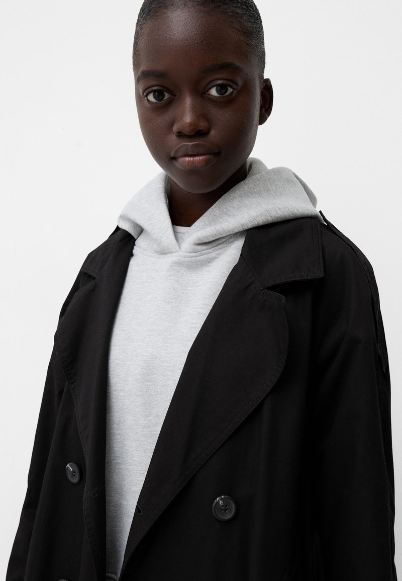 Młoda osoba o ciemnej karnacji ubrana w jasnoszary hoodie pod czarnym płaszczem zapinanym na guziki, patrząca prosto przed siebie na jednolitym tle.