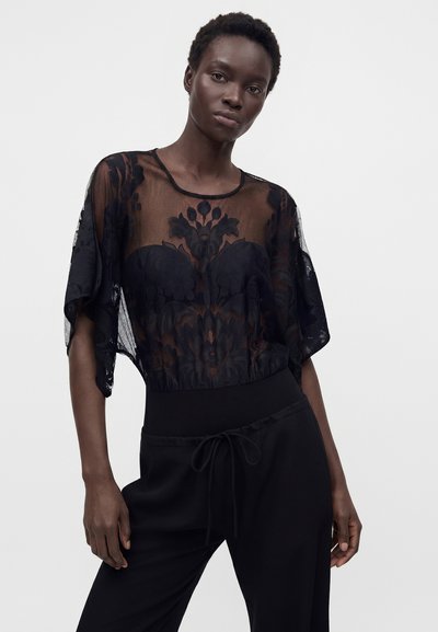 Femme portant un haut en dentelle noire transparente avec des motifs floraux sur un vêtement noir sans bretelles et un pantalon noir à cordon.