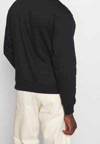 Sudadera negra con un ajuste relajado, puños y dobladillo acanalados. Combinada con pantalones de color claro, mostrando un diseño sencillo.