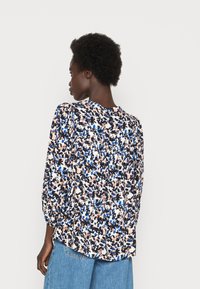 Blusa floral com mangas bufantes numa mistura de azul, preto e pêssego sobre um fundo branco. Tecido suave com um design fluido e decote redondo.