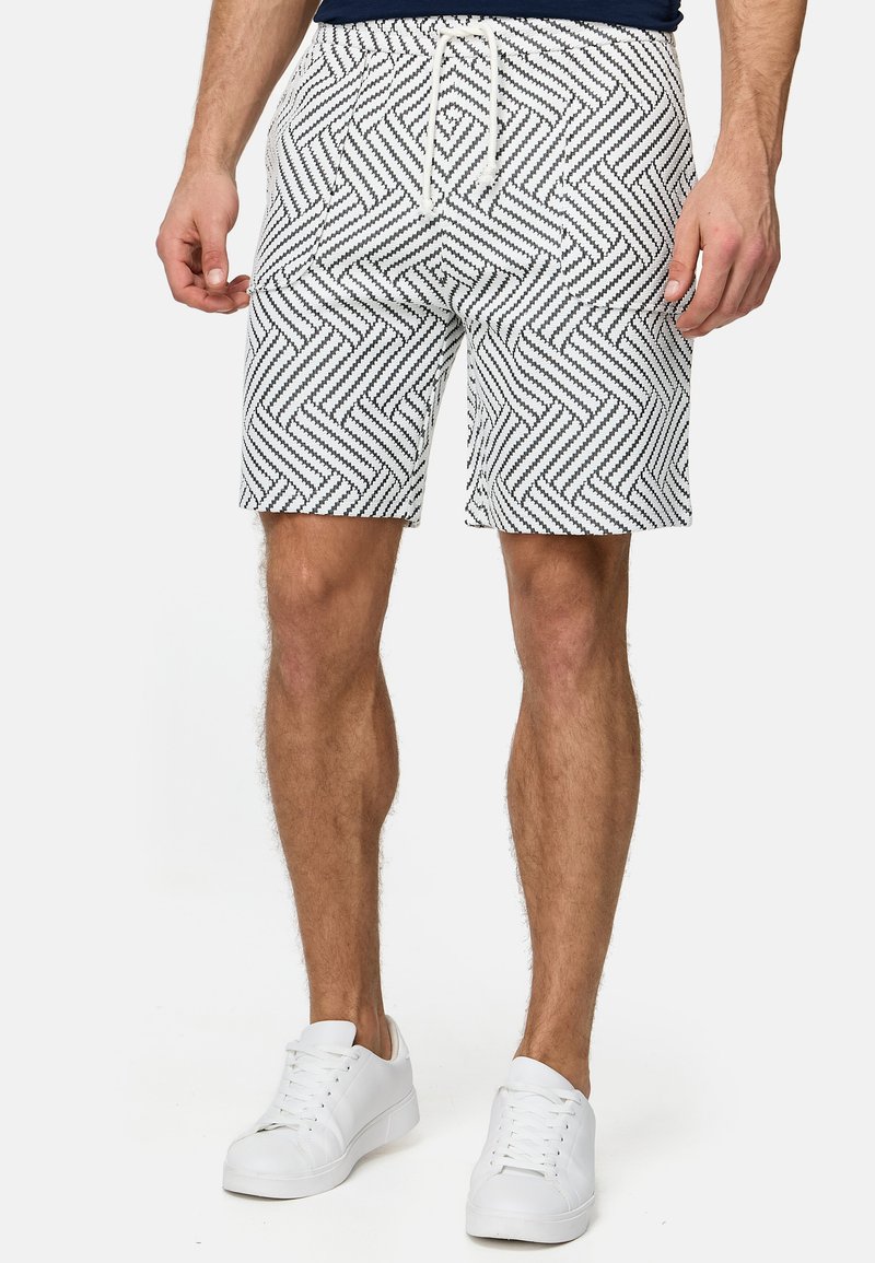 Bañador de hombre en tela blanca con un patrón geométrico negro. Cintura elástica con cordón. Se lleva con zapatillas blancas.