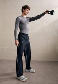 Grå langermet treningsgenser, svarte bukser med hvite striper, holder en svart kettlebell, iført grå joggesko. Jevn tekstur, tettsittende design.