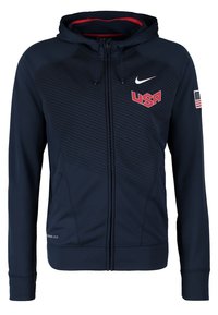 Sweat à capuche zippé bleu marine avec logo USA sur la poitrine, swoosh Nike au-dessus, drapeau américain sur la manche et doublure intérieure de la capuche rouge.