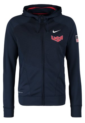 Mężczyzna ubrany w czarną bluzę z kapturem USA basketball, pasujące czarne i czerwone krótkie spodenki, białe skarpetki oraz białe buty sportowe idący do przodu.