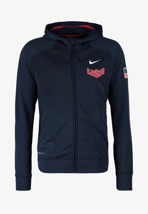 Homme portant un sweat à capuche noir USA basketball, un short noir et rouge assorti, des chaussettes blanches et des chaussures de sport blanches marchant vers l'avant.