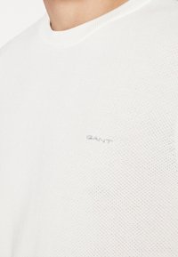 Gros plan sur une personne portant un pull en maille blanche texturée GANT à col rond avec le logo brodé sur la poitrine.