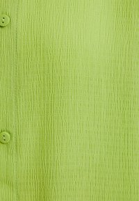 Tessuto verde chiaro testurizzato con un motivo a coste, dotato di chiusura con bottone. Il materiale ha una finitura leggermente lucida.