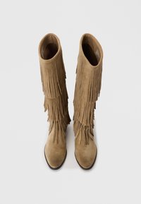 Bottes en daim marron jusqu'aux genoux avec des franges sur les côtés, bout pointu et texture lisse. L'intérieur est dépouillé.