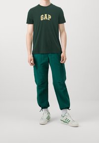 Persona vestida con una camiseta de GAP verde oscuro, pantalones cargo verdes con puños de cordón, calcetines blancos y zapatillas blancas con rayas verdes.