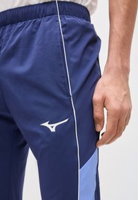 Άνδρας που φοράει αθλητικό παντελόνι σε ναυτικό μπλε με ανοιχτόχρωμες μπλε πλευρικές λωρίδες, λευκή περιμετρική ραφή, ελαστική μέση και λευκό λογότυπο Mizuno κοντά στην τσέπη.
