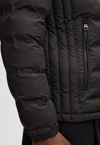 Piumino nero, caratterizzato da texture trapuntata, polsini a coste e una tasca laterale con cerniera e dettagli a contrasto.