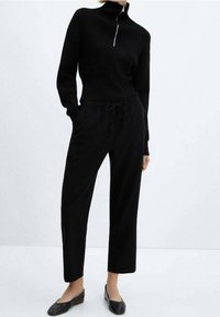 Sort strikket sweater med høj hals og sølv lynlås, parret med løse sorte bukser med snøre og flade sorte sko. Enkel, minimal design.