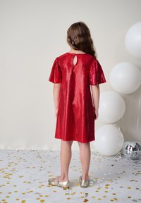 Rød palietkjole med korte puffærmer og en rund nøglehulssnit bagpå. Bærtes med guld sko mod en baggrund af konfetti og balloner.