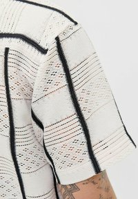 Haut en tissu texturé blanc avec des motifs ajourés et une bordure noire le long des bords. Manches courtes et détails de couture complexes présents.