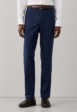 Man draagt marineblauwe nette broek, bruine leren riem, wit overhemd en donkerbruine loafers, staand voor een effen achtergrond.