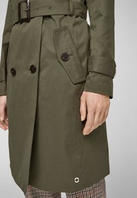 Trench coat khaki con bottoni doppi, ampie tasche laterali e vita con cintura. Caratterizzato da un dettaglio in grommet argentato all'orlo.