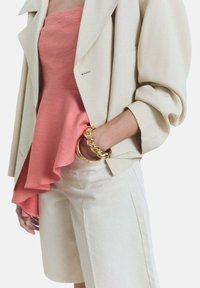 Blazer oversize beige abbinato a un top peplum corallo e pantaloni beige. Un braccialetto di catena dorata aggiunge un tocco all'outfit. Tessuto liscio e strutturato.