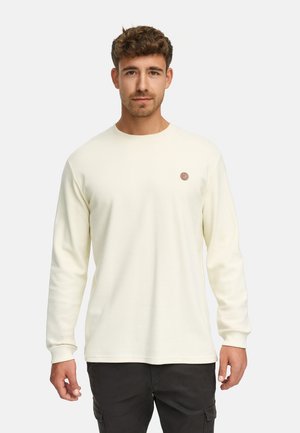 Man met kort bruin haar die een crème geribbeld shirt met lange mouwen en een zwarte broek draagt tegen een effen lichte achtergrond.