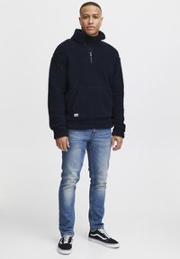 Mørkeblå fleece-pullover med halv glidelås, kenguru-lomme og teksturert overflate. Brukt med lyseblå jeans og svarte joggesko.