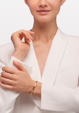 Kobieta w białej marynarce delikatnie trzyma nadgarstek ozdobiony cienką złotą bransoletką na jednolitym tle.