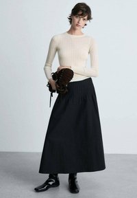 Cremfarbener gestreifter Langarmoberteil, kombiniert mit einem schwarzen, plissierten Maxirock. Das Modell hält ein braunes Schnür-Accessoire und trägt schwarze Stiefeletten.