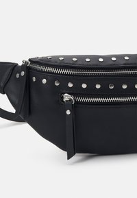 Sac banane en cuir noir avec des clous argentés et deux compartiments zippés avec des tirettes en cuir sur fond blanc.