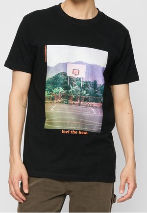 Homme portant un t-shirt noir avec un terrain de basket et un panier devant des collines et montagnes vertes, texte « ressentez la chaleur » en orange.