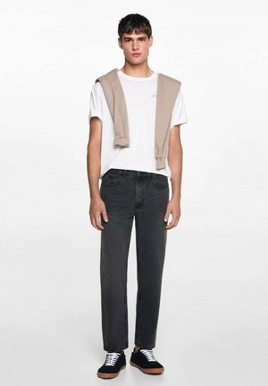 T-shirt bianco in cotone, maglione beige appoggiato sulle spalle, jeans grigio scuro e sneakers nere con suole marroni. Outfit semplice e informale.