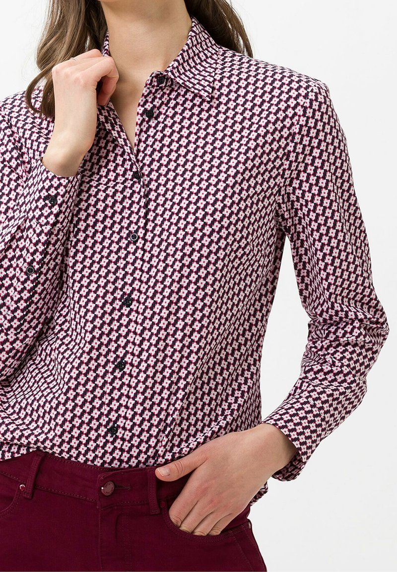 Chemise à manches longues avec boutons, arborant un motif géométrique rouge et blanc, avec un col pointu et des boutons noirs, confectionnée dans un tissu lisse.