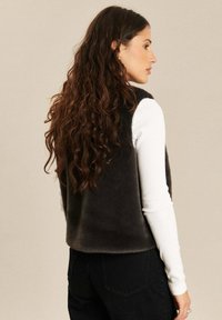 Vrouw met lang krullend haar, gekleed in een zwarte fuzzy vest over een witte lange mouwen top en zwarte broek, die zijwaarts kijkt tegen een beige achtergrond.