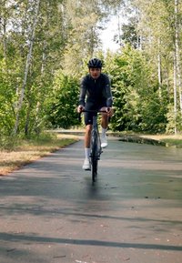 Cyklist på en svart landsvägscykel med smala däck, klädd i en grå cykeltröja och svarta shorts, cyklar längs en trädklädd stig.