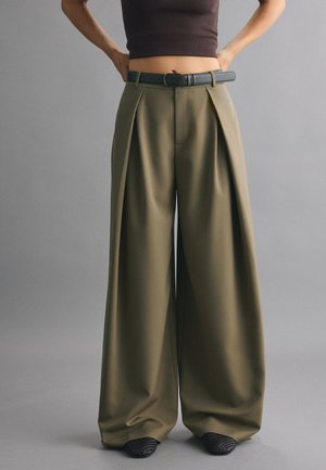 Personne portant un pantalon large taille haute vert olive avec des plis et une ceinture noire, associé à des chaussures noires texturées.