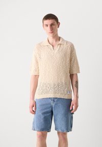 Beige strikket polo-skjorte med korte ermer og åpen vevstruktur, kombinert med blå denim-sjorts. Har en avslappet passform og krage.