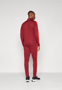 Homme de dos, portant un survêtement bordeaux avec des rayures rouges le long des manches et des jambes de pantalon, et des baskets noires avec des semelles blanches.