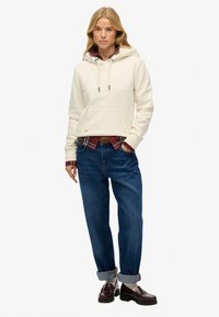 Sudadera crema con un bolsillo delantero y cordones, superpuesta sobre una camisa de cuadros roja y negra, combinada con unos vaqueros de corte relajado en azul y mocasines.