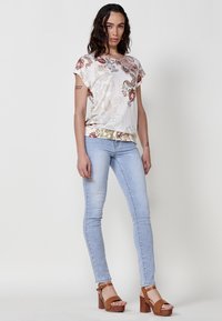 Haut blanc avec des motifs floraux et paisley, manches courtes et ourlet texturé ; associé à un jean skinny bleu clair et des sandales à talons bruns.