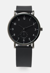 Montre analogique noire avec un cadran rond, des index horaires blancs et un sous-cadran. Dotée d'un bracelet en silicone noir mat et d'accents en métal.