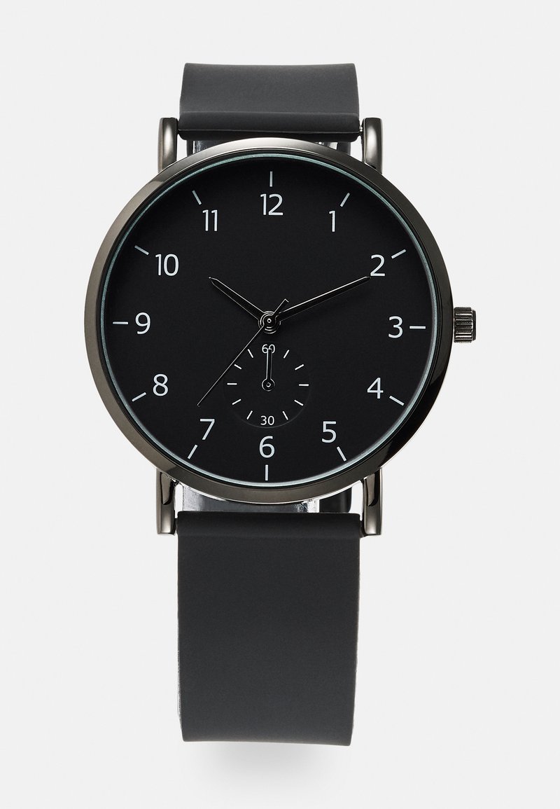 Montre analogique noire avec un cadran rond, des index horaires blancs et un sous-cadran. Dotée d'un bracelet en silicone noir mat et d'accents en métal.