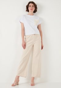 White cropped t-shirt with a scalloped beige pocket, paired with beige wide-leg pants, and light beige block-heeled sandals. Textured fabric.