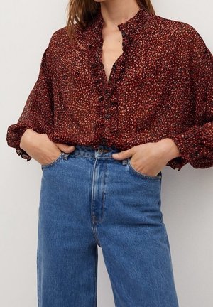 Kobieta ubrana w czerwoną bluzkę w wzory z falbankami oraz niebieskie dżinsy z wysokim stanem, ręce włożone do przednich kieszeni, na jednolitym tle.