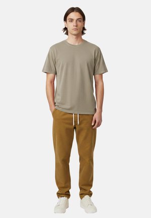 Giovane uomo in piedi, che indossa una maglietta a maniche corte color talpa, pantaloni marroni con coulisse e scarpe da ginnastica bianche, su sfondo semplice.