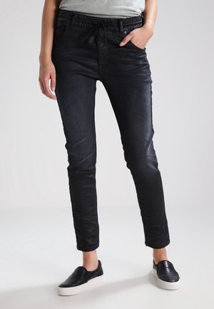Personne portant un pantalon noir slim avec une ceinture élastique, un cordon blanc et des chaussures noires sans lacets avec des semelles blanches.