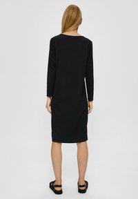 Schwarzes langärmliges Kleid aus strukturiertem Stoff, knielang, mit rundem Ausschnitt und geradem Schnitt, kombiniert mit schwarzen Sandalen.