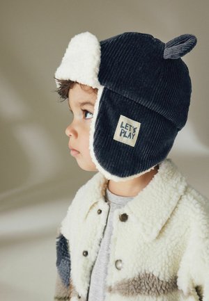 Marine corduroy muts met witte nepbont voering en oorflappen, voorzien van een grijze stoffen ooraccent en een "Laten we spelen" label. Kind dat een pluizige crèmekleurige jas draagt.